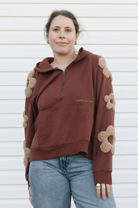 A woman wearing a brown half-zip hoodie with beige flower appliqués on the sleeves and shoulders, paired with blue jeans, standing against a white siding background.