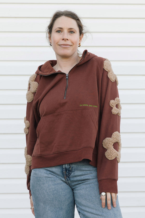 A woman wearing a brown half-zip hoodie with beige flower appliqués on the sleeves and shoulders, paired with blue jeans, standing against a white siding background.