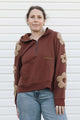 A woman wearing a brown half-zip hoodie with beige flower appliqués on the sleeves and shoulders, paired with blue jeans, standing against a white siding background.