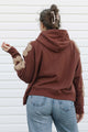 A person viewed from behind wearing a rust-brown hoodie with beige floral applique on the sleeves, paired with light-wash blue jeans, standing against a white siding background.
