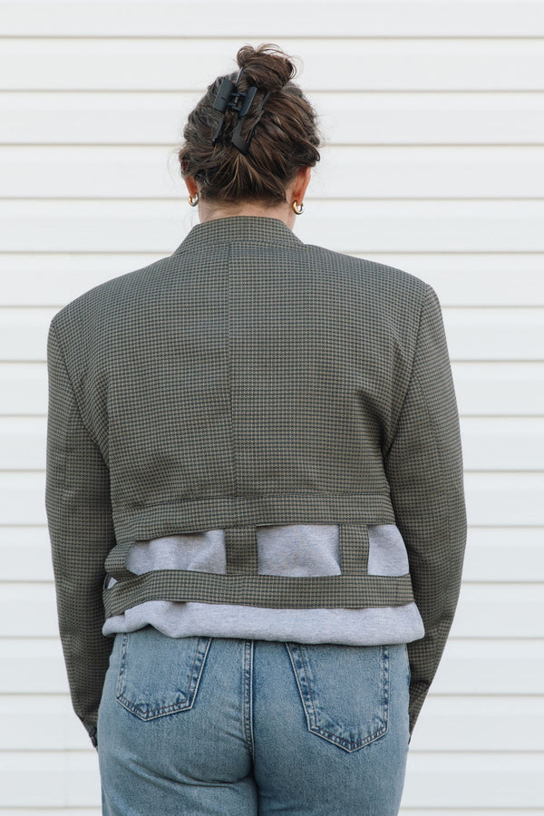 Person wearing a green checkered jacket over a gray shirt with blue jeans, standing against a white wall.