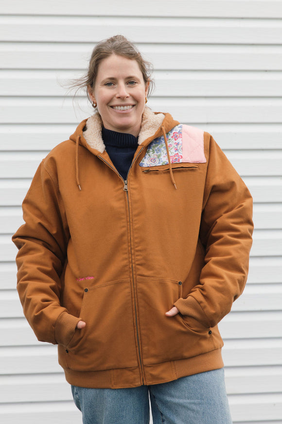 A woman wearing a rust-brown hooded jacket with a pink and blue floral patterned accent on the chest, standing in front of a white siding wall.
