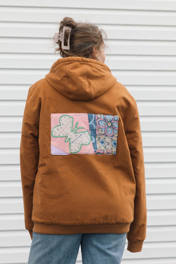 A person viewed from behind wearing a rust-brown hooded jacket with a colorful patchwork quilt design on the back, paired with blue jeans, standing against a white siding wall.
