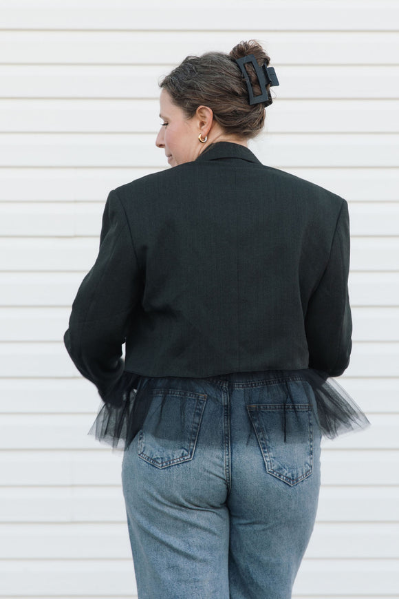 Person wearing a black blazer with tulle details and blue jeans against a white background