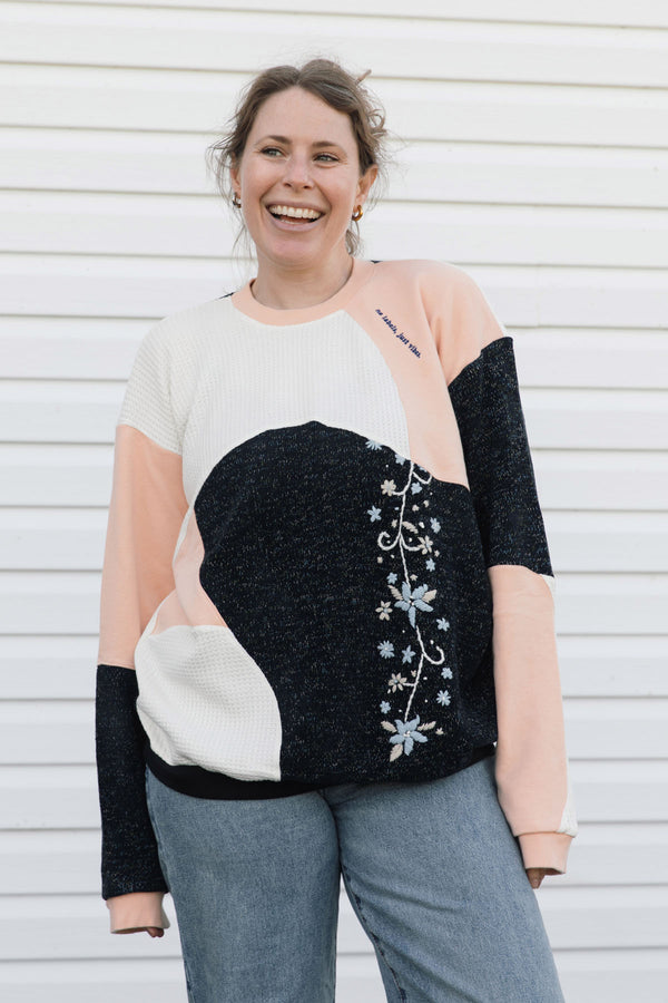 A woman smiling while wearing a color-block knit sweater with black, peach, and cream panels, featuring a floral vine pattern on the front, paired with blue jeans against a white siding background.