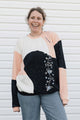 A woman smiling while wearing a color-block knit sweater with black, peach, and cream panels, featuring a floral vine pattern on the front, paired with blue jeans against a white siding background.
