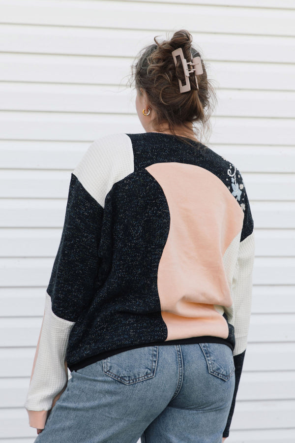 A woman viewed from behind wearing a color-block knit sweater with black, cream, and peach panels, paired with blue jeans against a white siding background.
