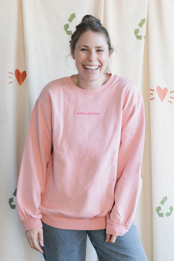 Person wearing a pink sweatshirt with 'no labels, just vibes.' embroidered on front, 100% cotton, upcycled in canada