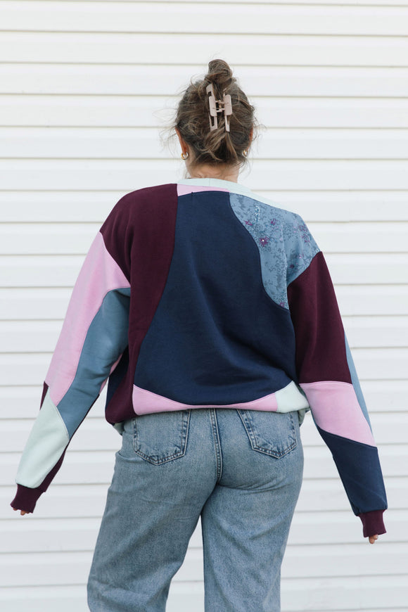 A person viewed from behind wearing a color-block patchwork sweatshirt with navy, burgundy, pink, and light blue panels, paired with light-wash jeans against a white siding background.