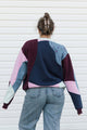 A person viewed from behind wearing a color-block patchwork sweatshirt with navy, burgundy, pink, and light blue panels, paired with light-wash jeans against a white siding background.