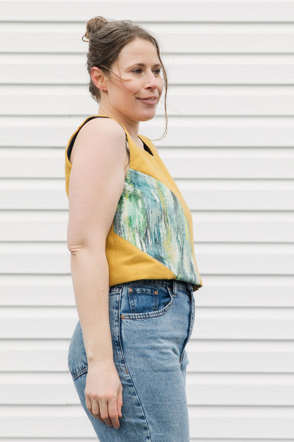 Woman wearing a mustard yellow sleeveless top with a colorful design and blue jeans against a white background
