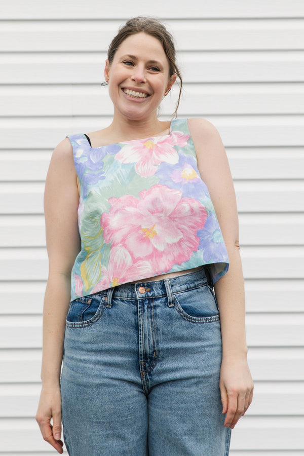 Woman wearing a floral sleeveless top and jeans standing against a white wall.