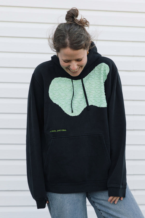 A woman wearing a black pullover hoodie with a large mint green textured heart graphic on the chest, paired with light blue jeans, standing against a white siding wall.