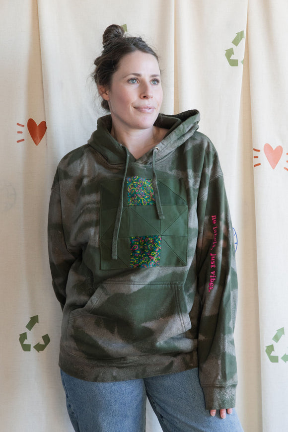 Person wearing a green camouflage hoodie with colorful design, with 'no labels, just vibes.' embroidered on sleeve, upcycled in canada