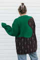 A person shown from the back wearing a chunky knit patchwork sweater with a bright green yoke and dark brown vertically striped body, standing against a white siding wall.
