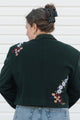 Person wearing a dark green jacket with floral embroidery on a white wall background