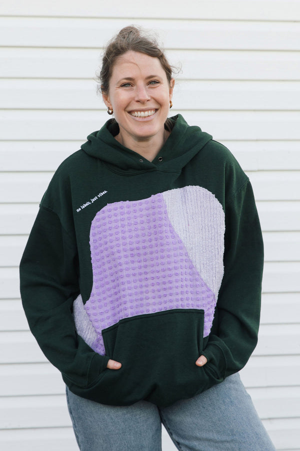 A woman smiling while wearing a dark green hoodie with a large lavender and light purple textured patch on the front, paired with gray jeans, standing against a white siding wall.