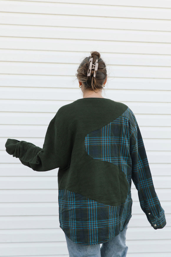 A person viewed from behind wearing a patchwork knit sweater with dark green ribbed sections and a dark green plaid panel with blue and orange lines, paired with light blue jeans against a white horizontal siding background.