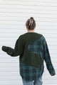 A person viewed from behind wearing a patchwork knit sweater with dark green ribbed sections and a dark green plaid panel with blue and orange lines, paired with light blue jeans against a white horizontal siding background.