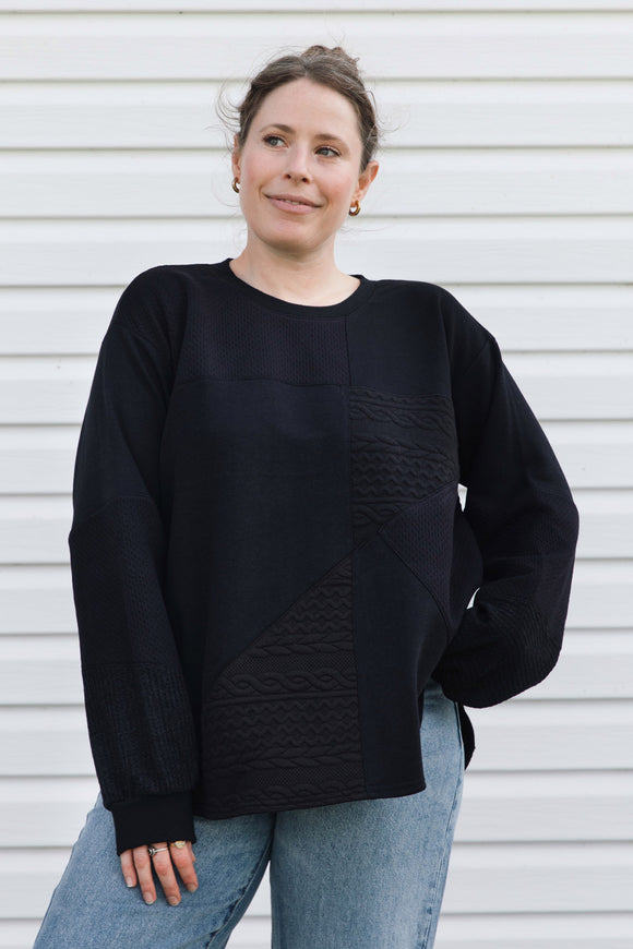 A woman wearing a black long-sleeve knit sweater with textured geometric paneling on the front, paired with light blue jeans, standing against a white horizontal siding wall.