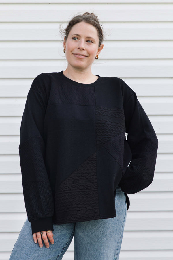 A woman wearing a black long-sleeve knit sweater with textured geometric paneling on the front, paired with light blue jeans, standing against a white horizontal siding wall.