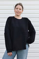 A woman wearing a black long-sleeve knit sweater with textured geometric paneling on the front, paired with light blue jeans, standing against a white horizontal siding wall.