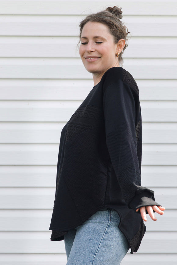 A woman wearing a black long-sleeve knit top with textured lace-like detailing on the chest and cuffs, paired with light blue jeans, standing against a white horizontal siding wall.