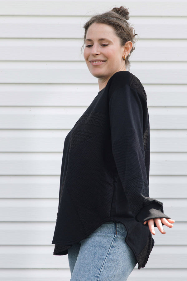 A woman wearing a black long-sleeve knit top with textured lace-like detailing on the chest and cuffs, paired with light blue jeans, standing against a white horizontal siding wall.