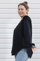 A woman wearing a black long-sleeve knit top with textured lace-like detailing on the chest and cuffs, paired with light blue jeans, standing against a white horizontal siding wall.