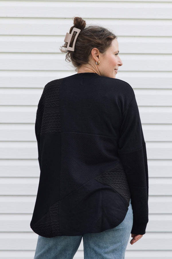 A woman wearing a black knit sweater with textured knit panel details on the back, shown from behind against a white siding background.