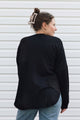 A woman wearing a black knit sweater with textured knit panel details on the back, shown from behind against a white siding background.