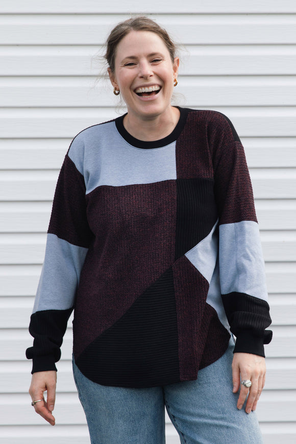 A woman smiling and laughing while wearing a color-block knit sweater with dark burgundy, light blue, and black panels, paired with blue jeans, standing in front of a white horizontal siding wall.