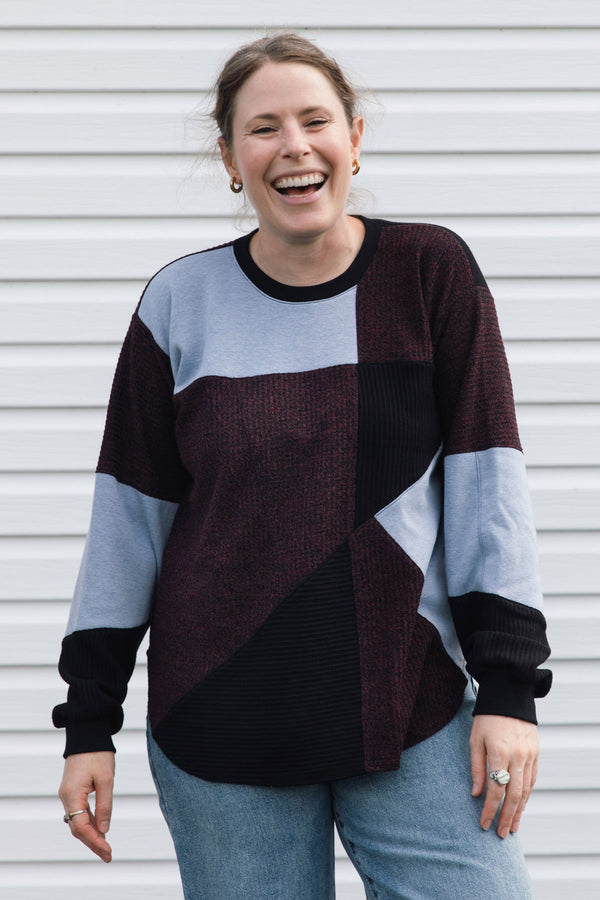 A woman smiling and laughing while wearing a color-block knit sweater with dark burgundy, light blue, and black panels, paired with blue jeans, standing in front of a white horizontal siding wall.