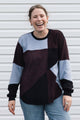 A woman smiling and laughing while wearing a color-block knit sweater with dark burgundy, light blue, and black panels, paired with blue jeans, standing in front of a white horizontal siding wall.