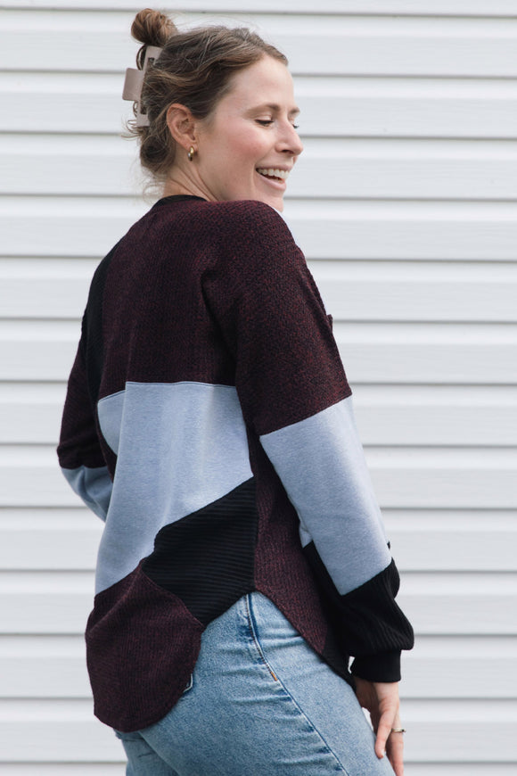 A woman wearing a color-block knit sweater in burgundy, light blue, and black, paired with light-wash blue jeans, standing in front of a white horizontal-slat wall.