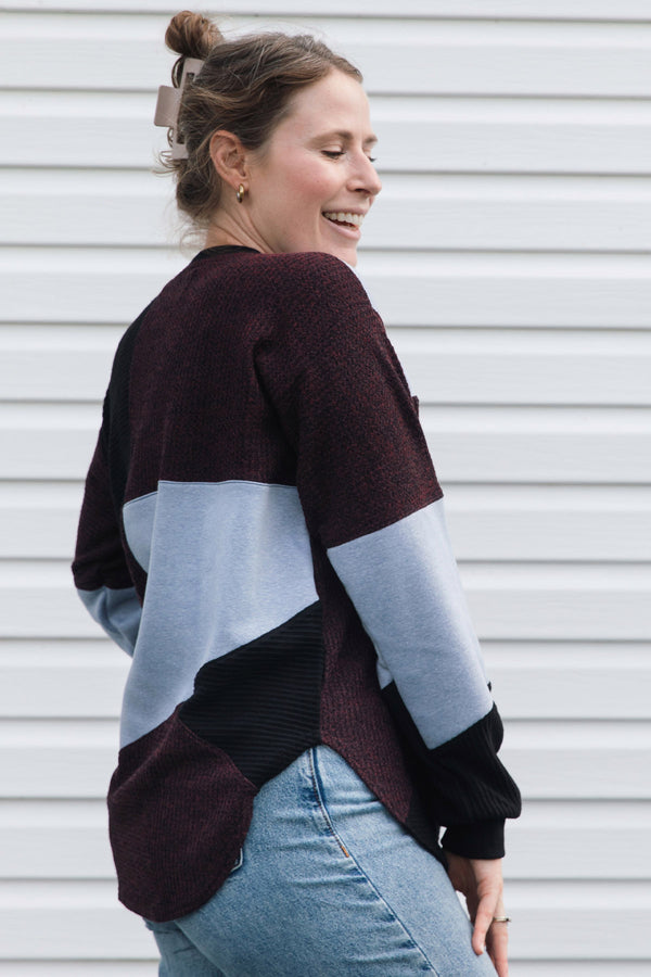 A woman wearing a color-block knit sweater in burgundy, light blue, and black, paired with light-wash blue jeans, standing in front of a white horizontal-slat wall.