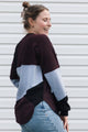 A woman wearing a color-block knit sweater in burgundy, light blue, and black, paired with light-wash blue jeans, standing in front of a white horizontal-slat wall.