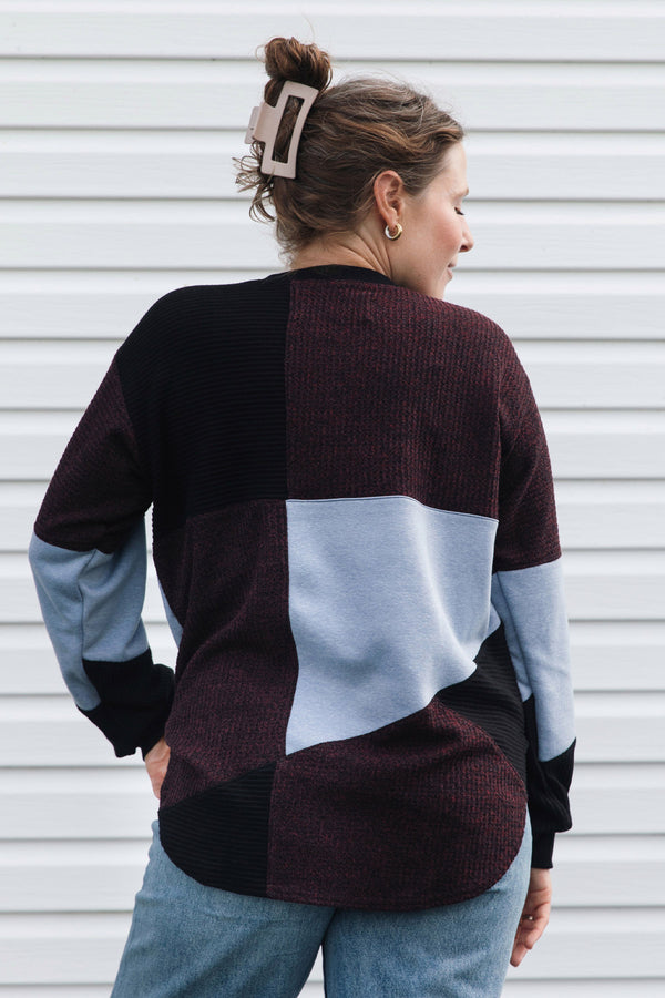 A woman shown from the back wearing a color-block knit sweater with panels of dark burgundy, black, light blue, and gray, paired with blue jeans against a white siding background.