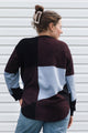 A woman shown from the back wearing a color-block knit sweater with panels of dark burgundy, black, light blue, and gray, paired with blue jeans against a white siding background.