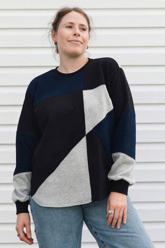 A woman wearing a color-block knit sweater with navy, black, and heather gray panels, paired with blue jeans, standing in front of a white siding wall.