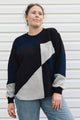 A woman wearing a color-block knit sweater with navy, black, and heather gray panels, paired with blue jeans, standing in front of a white siding wall.