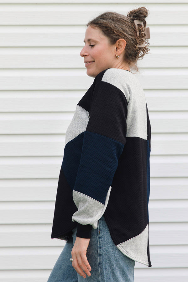 A woman wearing a color-block knit sweater in navy, light gray, and off-white, paired with blue jeans, standing in profile against a white horizontal-slat wall.