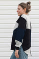 A woman wearing a color-block knit sweater in navy, light gray, and off-white, paired with blue jeans, standing in profile against a white horizontal-slat wall.
