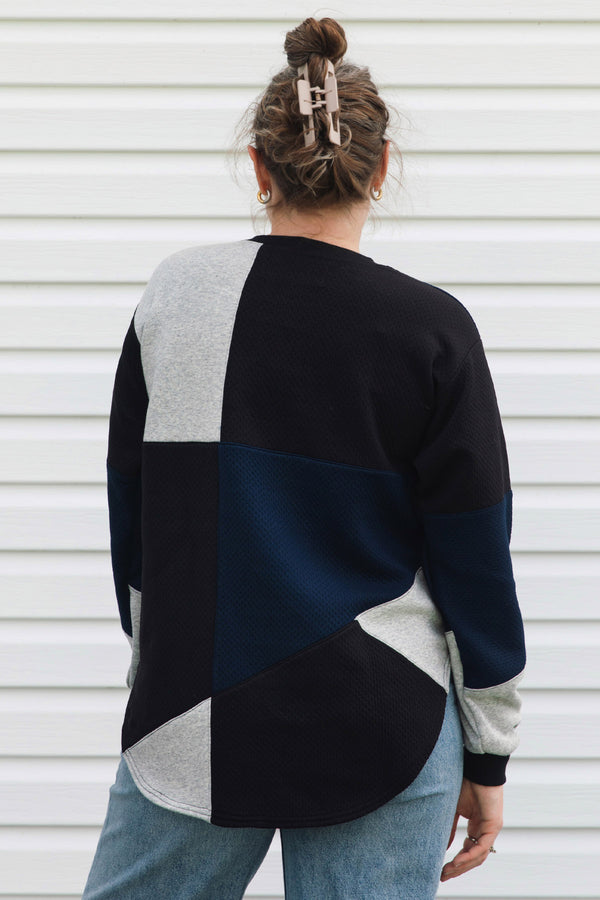 A woman viewed from behind wearing a color-block knit sweater with geometric panels in black, navy, and light heather gray, paired with light blue jeans against a white siding background.