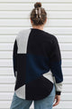 A woman viewed from behind wearing a color-block knit sweater with geometric panels in black, navy, and light heather gray, paired with light blue jeans against a white siding background.
