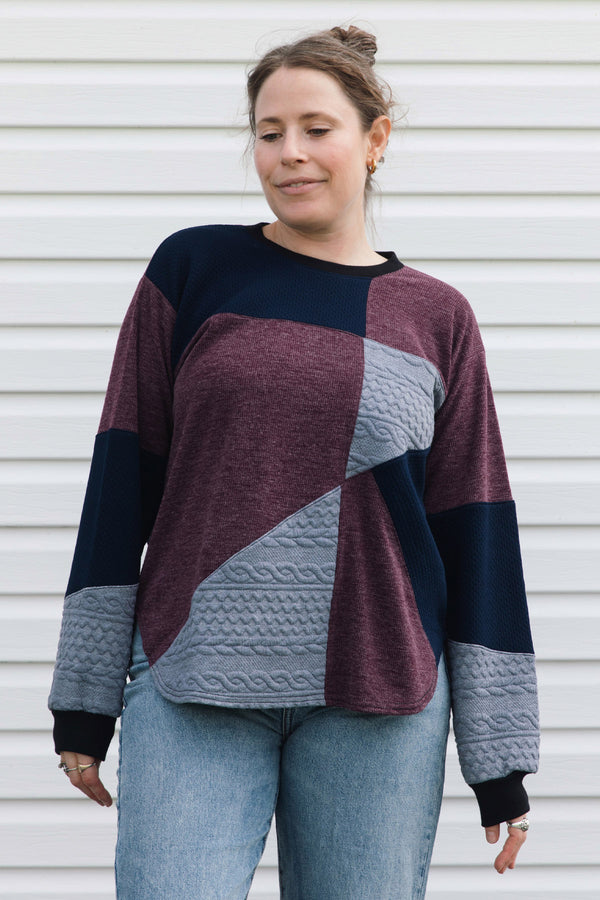 A woman wearing a color-block knit sweater with burgundy, navy, and gray panels, paired with light blue jeans, standing against a white siding background.