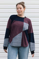 A woman wearing a color-block knit sweater with burgundy, navy, and gray panels, paired with light blue jeans, standing against a white siding background.