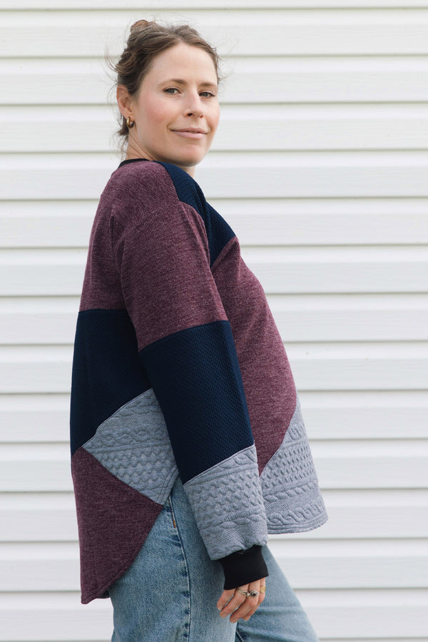 A woman wearing a color-block knit cardigan with burgundy, navy, and light gray panels, paired with blue jeans, standing against a white siding background.
