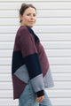 A woman wearing a color-block knit cardigan with burgundy, navy, and light gray panels, paired with blue jeans, standing against a white siding background.