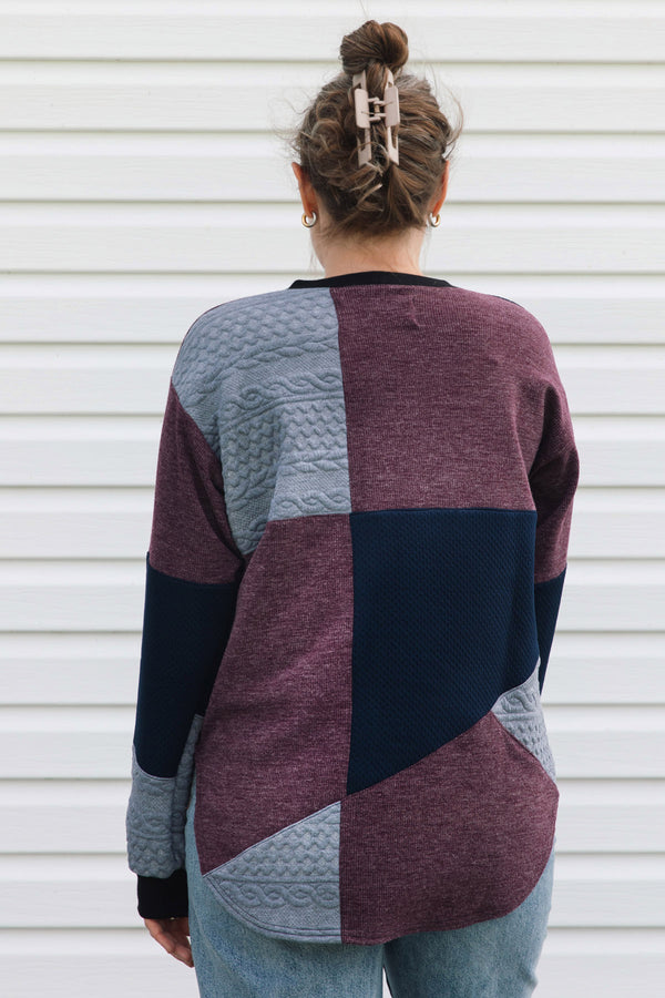A woman viewed from behind wearing a color-block knit sweater with panels of burgundy, navy, and light blue-gray, paired with light-wash jeans against a white siding background.